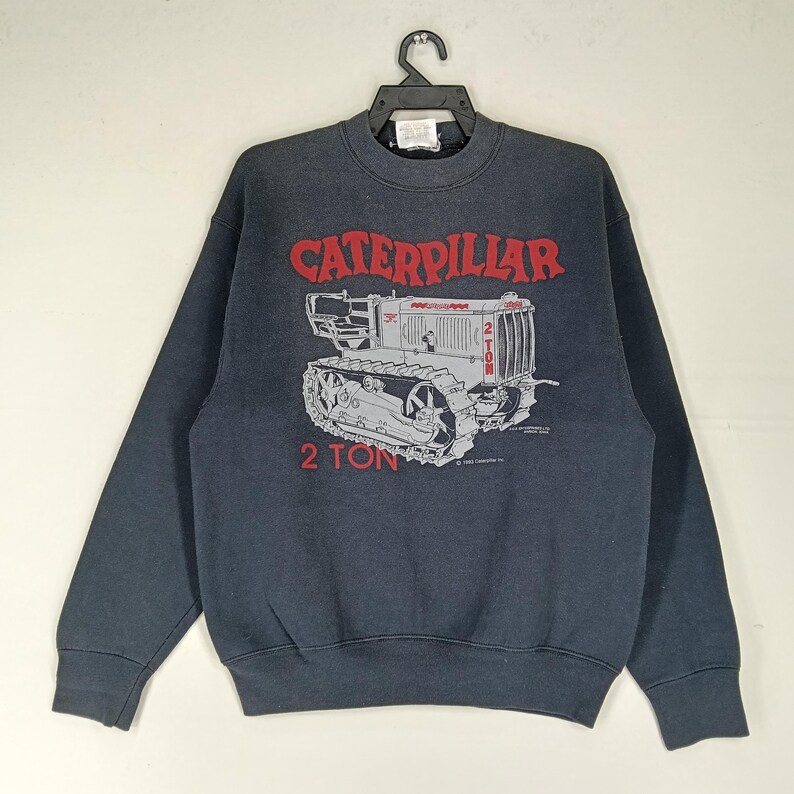 K&ouml;nnte beinhalten: Dunkelgraues Sweatshirt mit einem roten und grauen Caterpillar-Traktor-Motiv. Der Text "CATERPILLAR" steht dar&uuml;ber und "2 TON" darunter. Es hat einen Rundhalsausschnitt und lange &Auml;rmel.
