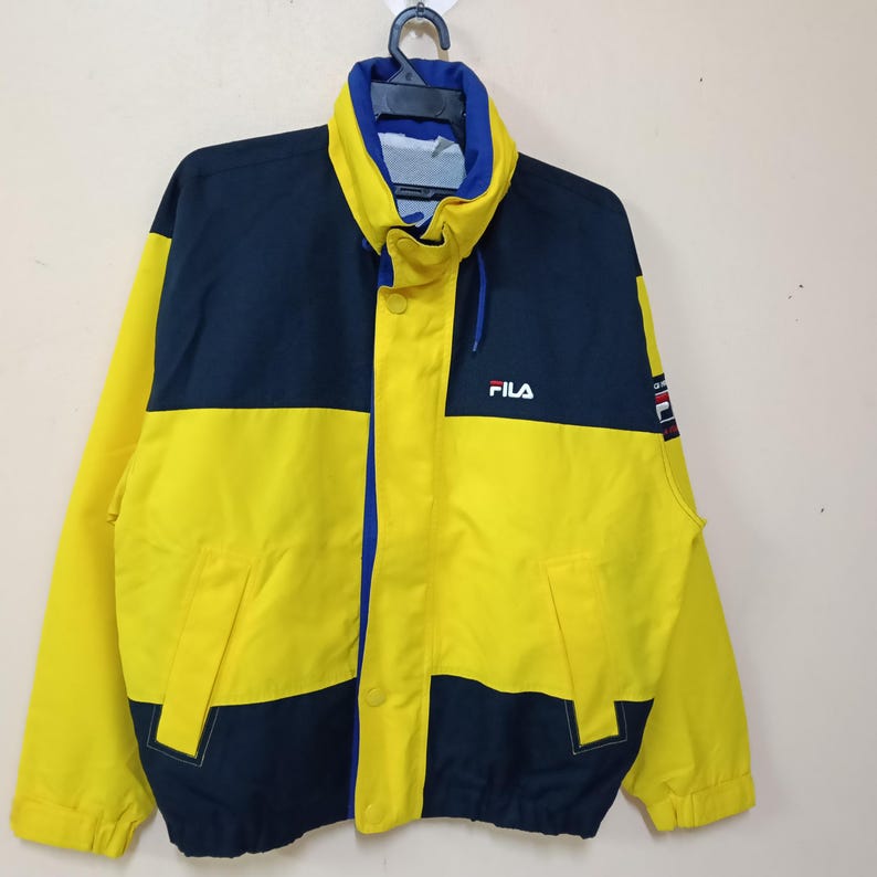 Puede incluir: Chaqueta FILA vintage en amarillo y azul marino. La chaqueta presenta un cuello alto, una cremallera frontal y el logotipo de FILA en el pecho. La chaqueta tiene un dise&ntilde;o de bloques de color con un esquema de color azul y amarillo.