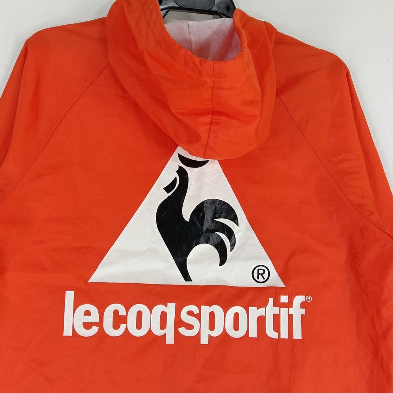 K&ouml;nnte beinhalten: Leuchtend orangefarbene Kapuzenjacke mit einem wei&szlig;en Dreieckslogo, das einen schwarzen Hahn zeigt. Der Markenname "le coq sportif" ist wei&szlig; aufgedruckt. Die Jacke hat eine Kapuze und ein wei&szlig;es Innenfutter.