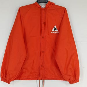 K&ouml;nnte beinhalten: Leuchtend orangefarbene Kapuzenjacke mit Knopfleiste. Die Jacke hat ein wei&szlig;es Dreieckslogo mit einem Hahn und dem Text "le coq sportif". Sie hat einen Kordelzug am Saum und ist aus leichtem Material gefertigt.