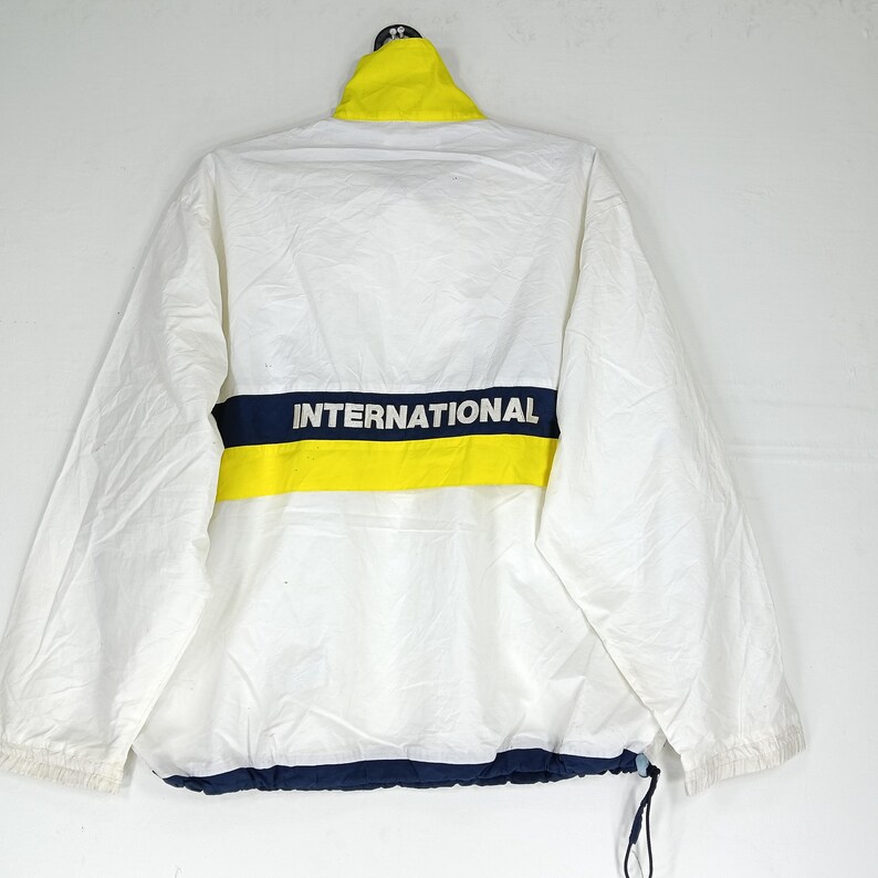 May include: White jacket with a yellow collar and a navy blue and yellow stripe across the back. The word "INTERNATIONAL" is printed in white on the navy blue stripe. The jacket has a drawstring at the bottom.