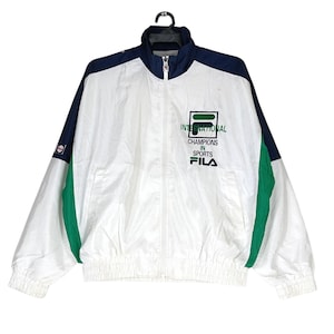 May include: White, navy blue, and green Fila track jacket with a full zipper closure. The jacket features the text "INTERNATIONAL CHAMPIONS IN SPORTS" and the Fila logo on the chest. The jacket has a stand-up collar and elastic cuffs and hem.