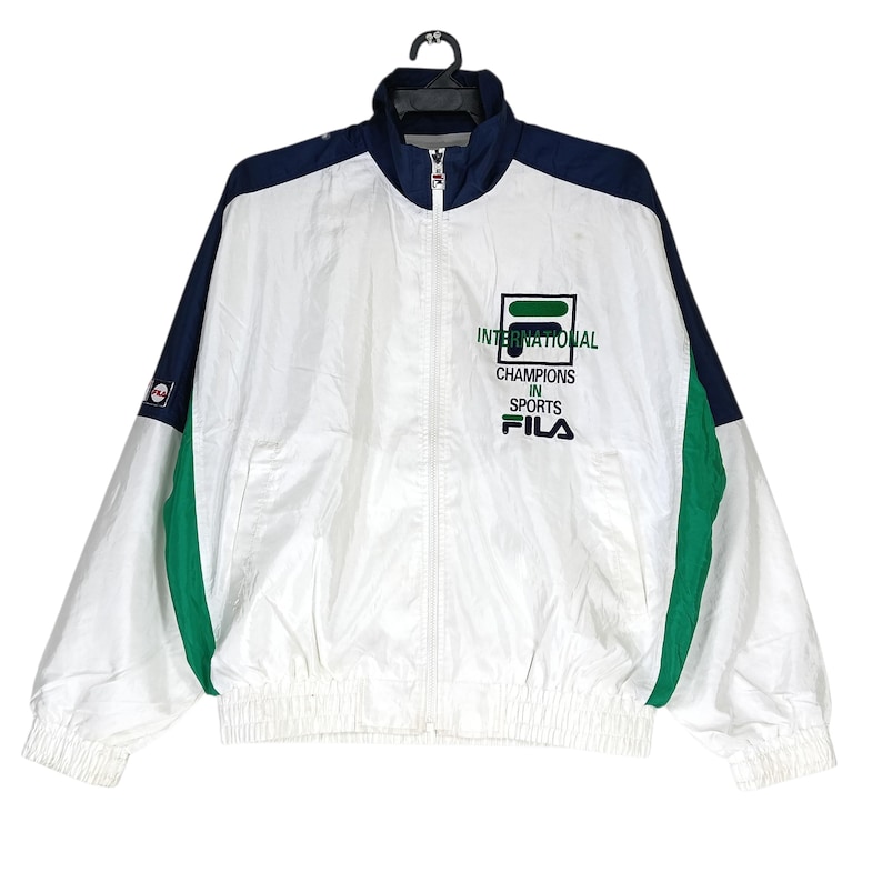 May include: White, navy blue, and green Fila track jacket with a full zipper closure. The jacket features the text "INTERNATIONAL CHAMPIONS IN SPORTS" and the Fila logo on the chest. The jacket has a stand-up collar and elastic cuffs and hem.