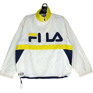 May include: White Fila jacket with a yellow and navy blue stripe across the chest. The jacket has a high collar and a half-zip closure. The Fila logo is prominently displayed in navy blue and yellow on the front.