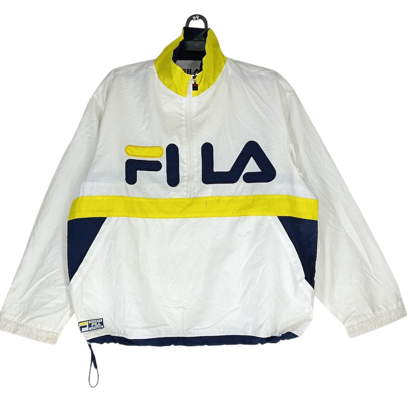 May include: White Fila jacket with a yellow and navy blue stripe across the chest. The jacket has a high collar and a half-zip closure. The Fila logo is prominently displayed in navy blue and yellow on the front.