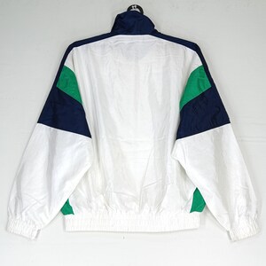 May include: White, navy blue, and green athletic jacket. The jacket has a high collar and a full-length zipper. The sleeves and body are white, with navy blue and green color blocks on the shoulders. The jacket has elastic cuffs and a drawstring hem.