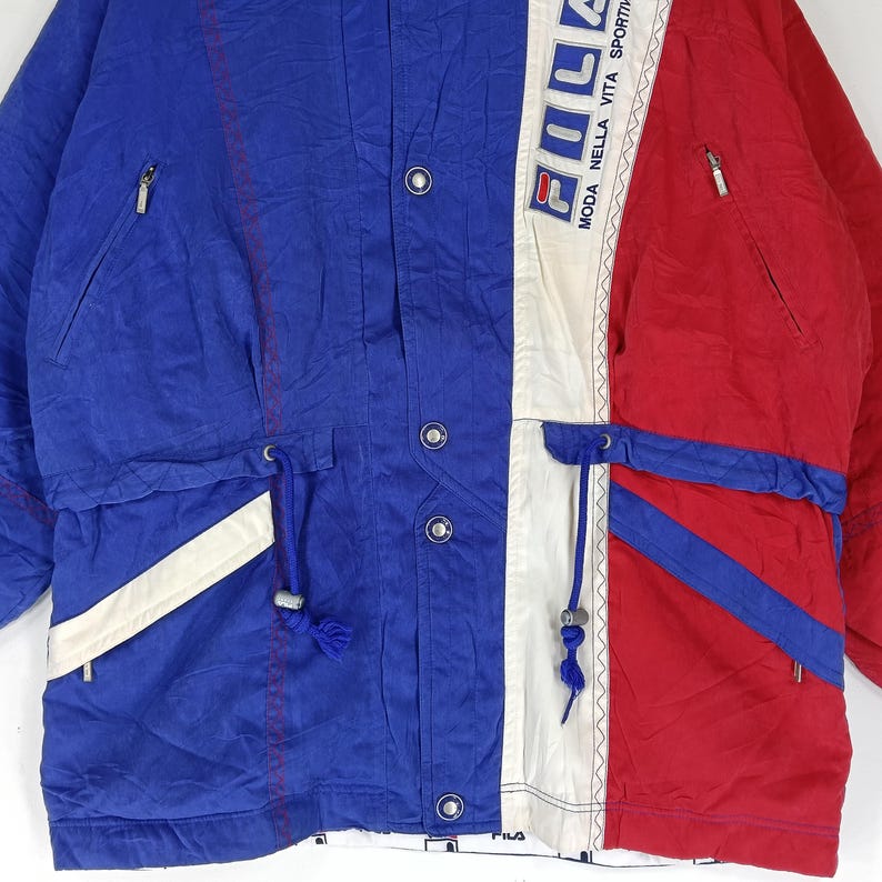 May include: A vintage color-blocked jacket in blue, white, and red. The jacket features a drawstring waist, multiple pockets, and the word "FILA" printed vertically on a white panel. The jacket has a sporty aesthetic.