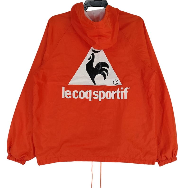 K&ouml;nnte beinhalten: Leuchtend orangefarbene Kapuzenjacke mit dem Le Coq Sportif Logo auf dem R&uuml;cken. Das Logo zeigt einen schwarzen Hahn in einem wei&szlig;en Dreieck, darunter der Markenname. Die Jacke hat einen Kordelzug am Saum und ein wei&szlig;es Futter in der Kapuze.