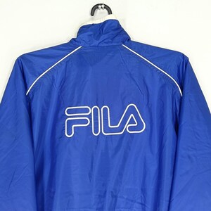 May include: A blue Fila jacket with a white collar and white trim. The Fila logo is prominently displayed in white on the back of the jacket. The jacket is hanging on a black hanger.