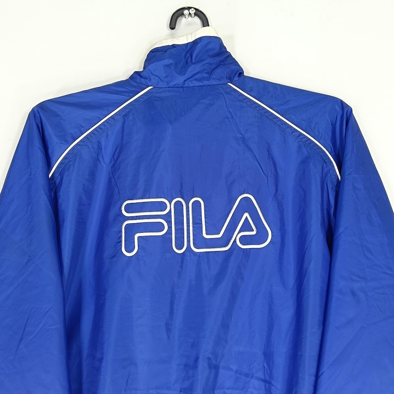 May include: A blue Fila jacket with a white collar and white trim. The Fila logo is prominently displayed in white on the back of the jacket. The jacket is hanging on a black hanger.