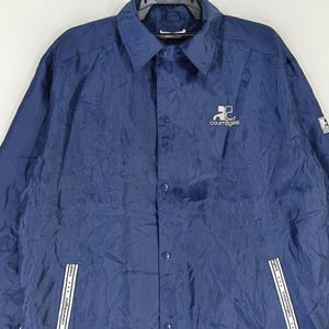 May include: A navy blue nylon jacket with a button-up front and a white logo that says "Courr&egrave;ges" on the left chest.