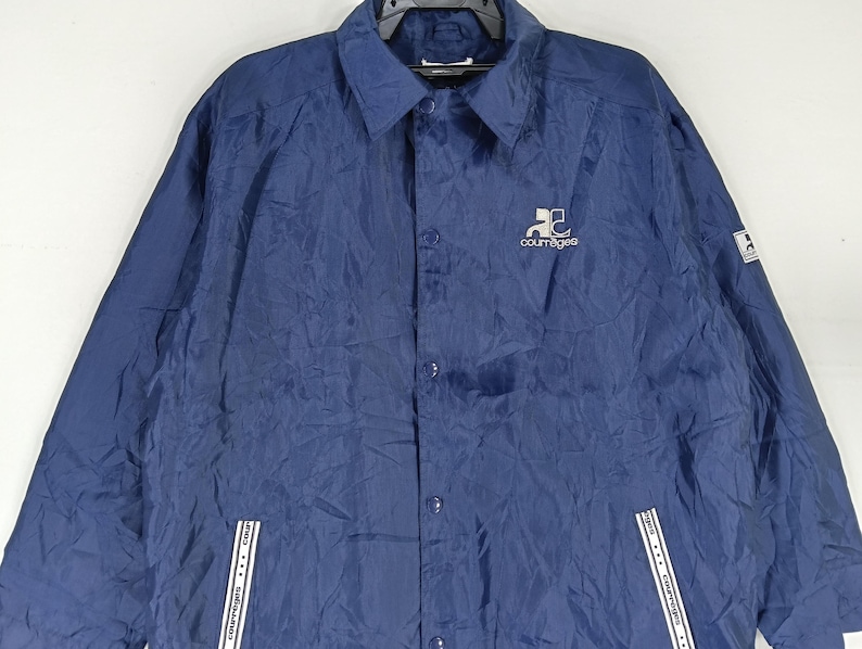 May include: A navy blue nylon jacket with a button-up front and a white logo that says "Courr&egrave;ges" on the left chest.