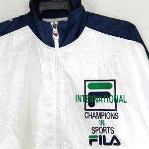 May include: White and navy blue FILA track jacket with a full zipper and a high collar. The jacket features the FILA logo and the words "INTERNATIONAL CHAMPIONS IN SPORTS" in green and navy blue.