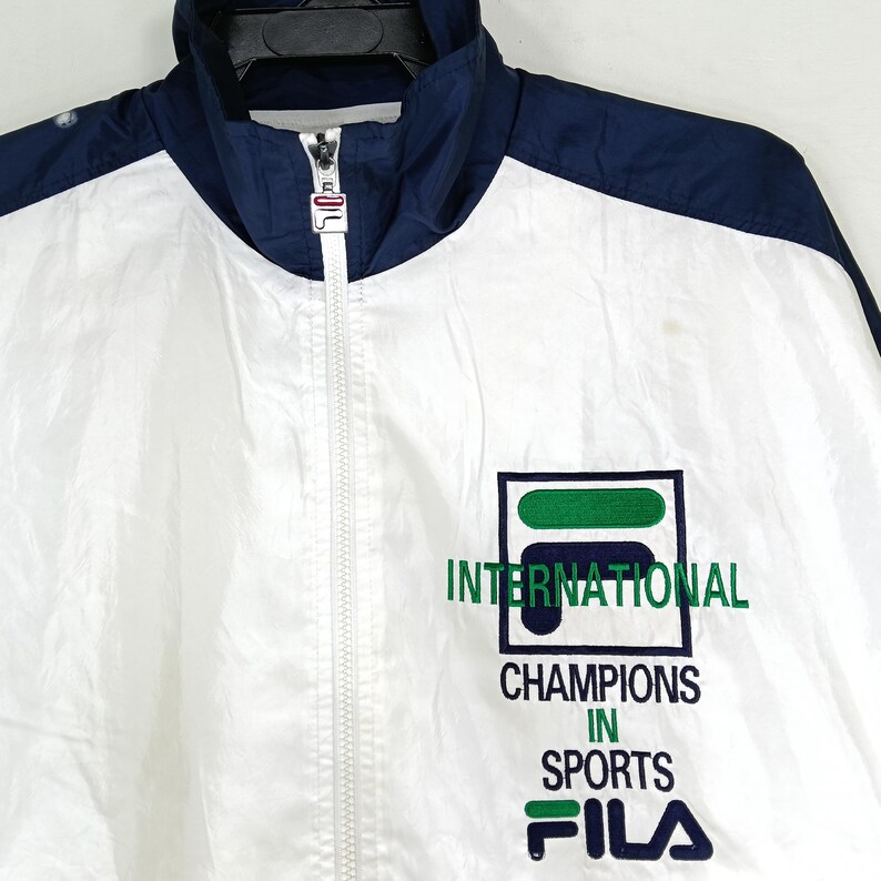 May include: White and navy blue FILA track jacket with a full zipper and a high collar. The jacket features the FILA logo and the words "INTERNATIONAL CHAMPIONS IN SPORTS" in green and navy blue.