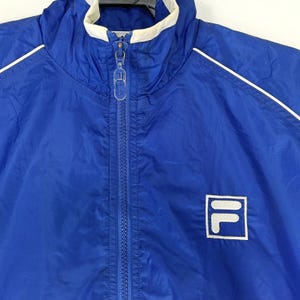 May include: A blue nylon jacket with a white collar and white trim along the shoulders. The jacket has a zipper closure and a small logo on the lower right. The jacket is a sporty style.
