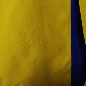 Puede incluir: Primer plano de una tela amarilla con una franja vertical azul en el lado derecho. La tela amarilla tiene un aspecto texturizado. Se ven peque&ntilde;as manchas oscuras en la tela amarilla.