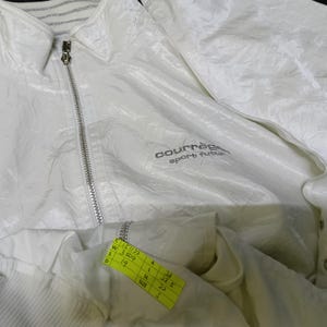 May include: A white Courr&egrave;ges sport futur jacket with a zipper closure and a collared neckline. The jacket has the brand name and logo on the front. A yellow tag with measurements is attached to the jacket.