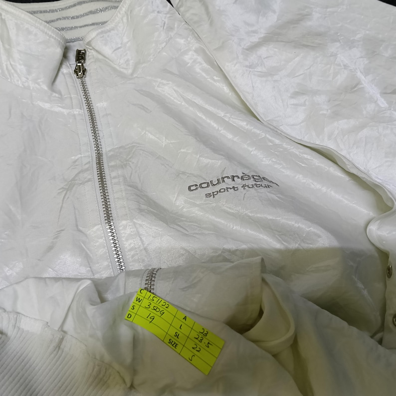 May include: A white Courr&egrave;ges sport futur jacket with a zipper closure and a collared neckline. The jacket has the brand name and logo on the front. A yellow tag with measurements is attached to the jacket.