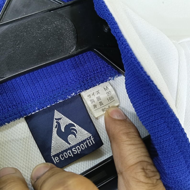May include: A white polo shirt with a blue trim and a Le Coq Sportif logo. The shirt has a size tag that reads "Size M, Chest 90 cm, Length 165 cm".