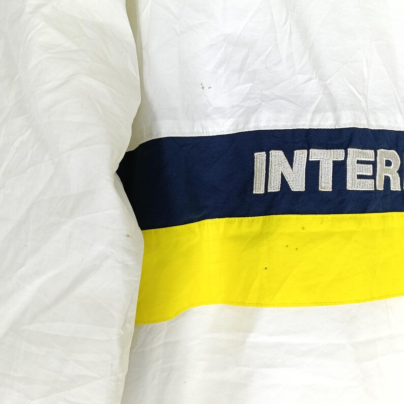 May include: White jacket with a navy blue and yellow stripe across the chest. The word "INTER" is embroidered in white on the navy stripe. The jacket appears to be made of a lightweight material.