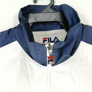 May include: A white and navy blue FILA jacket with a full zipper and a hood. The jacket has the FILA logo on the chest. The jacket is hanging on a black hanger.