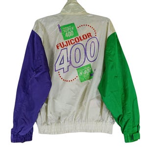 Puede incluir: Chaqueta de nailon vintage con cuerpo color crema, mangas moradas y manga derecha verde. La parte trasera presenta el texto "FUJICOLOR 400" en letras moradas grandes, con texto y gráficos verdes y morados más pequeños.