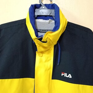 Puede incluir: Chaqueta negra y amarilla con cuello azul y logotipo "FILA" blanco. La chaqueta tiene un cord&oacute;n y cierre de bot&oacute;n. La chaqueta est&aacute; colgada de una percha negra.