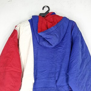 May include: A color-blocked jacket with a hood, featuring red, white, and blue panels. The jacket is hanging on a black hanger. The hood is red, and the body of the jacket is blue and white.
