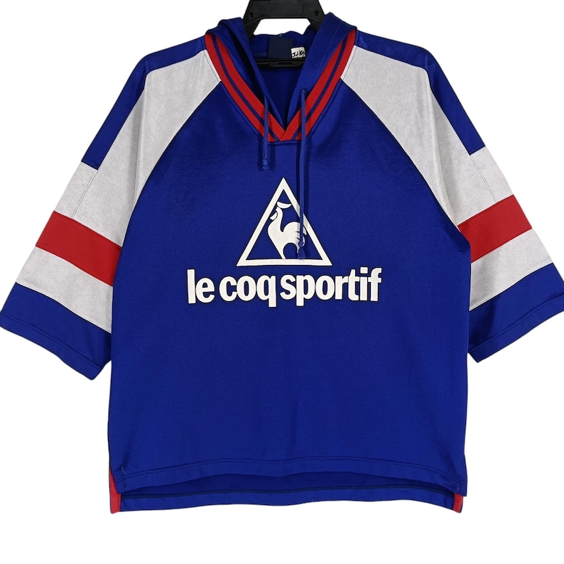 K&ouml;nnte beinhalten: Ein blaues, wei&szlig;es und rotes, kurz&auml;rmeliges Kapuzen-Sweatshirt mit dem Logo "le coq sportif" und einem Hahn in einem Dreieck.