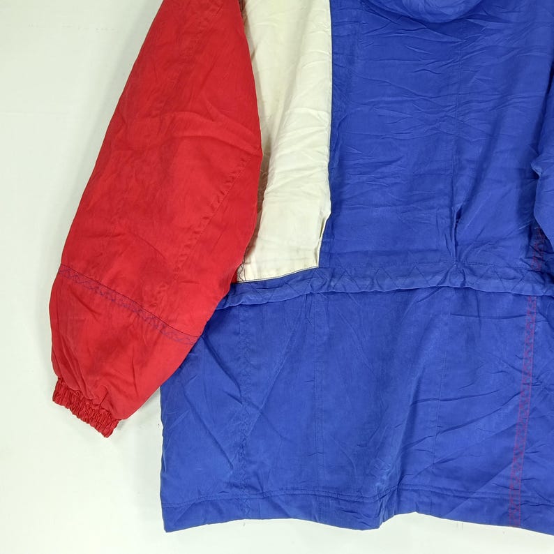 May include: A colorful jacket with a red sleeve, a white panel, and a blue body. The jacket has a hood and a zipper. The fabric appears to be a lightweight, possibly nylon material. The jacket is designed for outdoor wear.