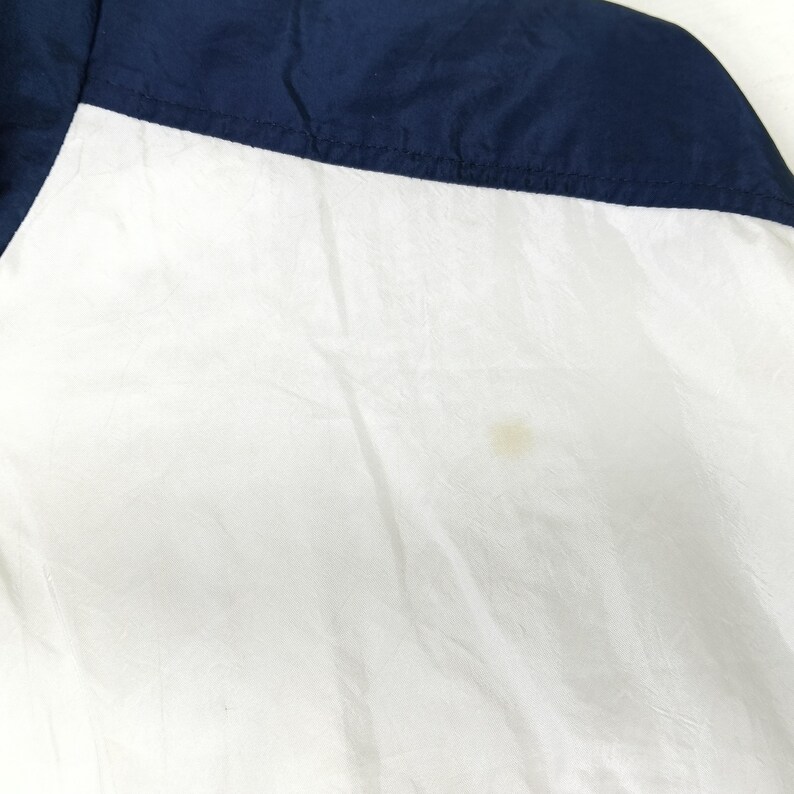 May include: A section of a white and navy blue jacket. The jacket is divided diagonally, with the top portion in navy blue and the bottom in white. The fabric appears to be lightweight and slightly wrinkled.