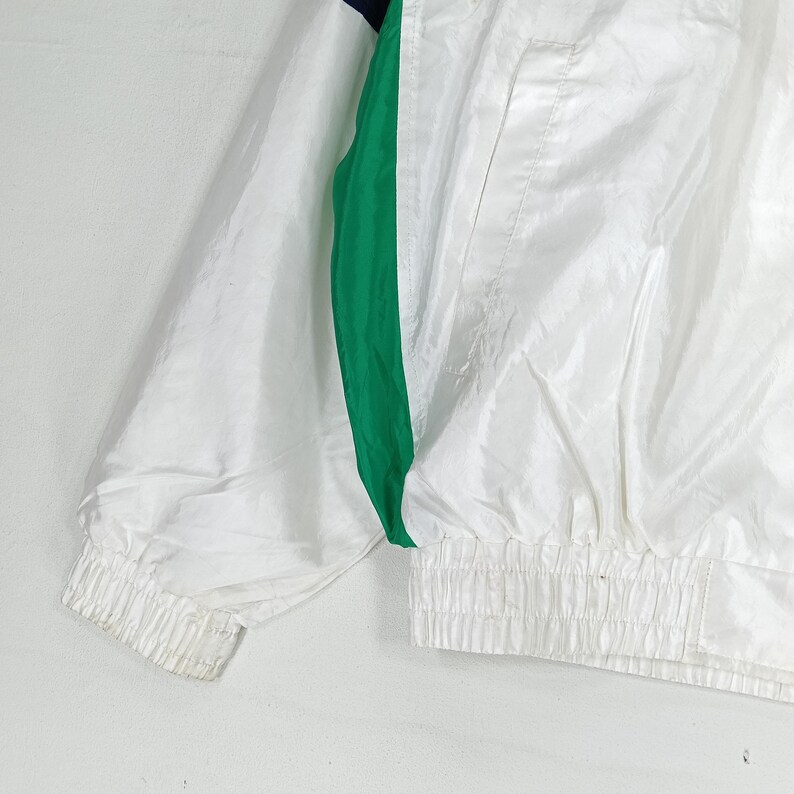 May include: White nylon jacket with a green and navy stripe down the side. The jacket has a ribbed cuff and a gathered waist. The jacket is made of a lightweight material.