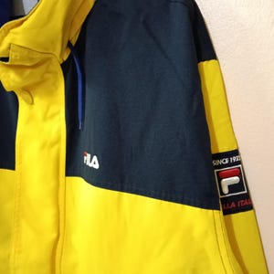 Puede incluir: Chaqueta amarilla y azul marino con el logo de Fila. La chaqueta tiene un cord&oacute;n azul y las palabras "SINCE 1923" e "ILLA ITAL" son visibles en un parche.