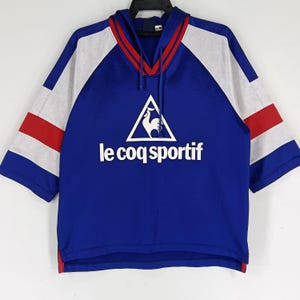K&ouml;nnte beinhalten: Ein blaues, wei&szlig;es und rotes Kurzarm-Kapuzenpullover mit dem Le Coq Sportif Logo und einem Hahn-Design.
