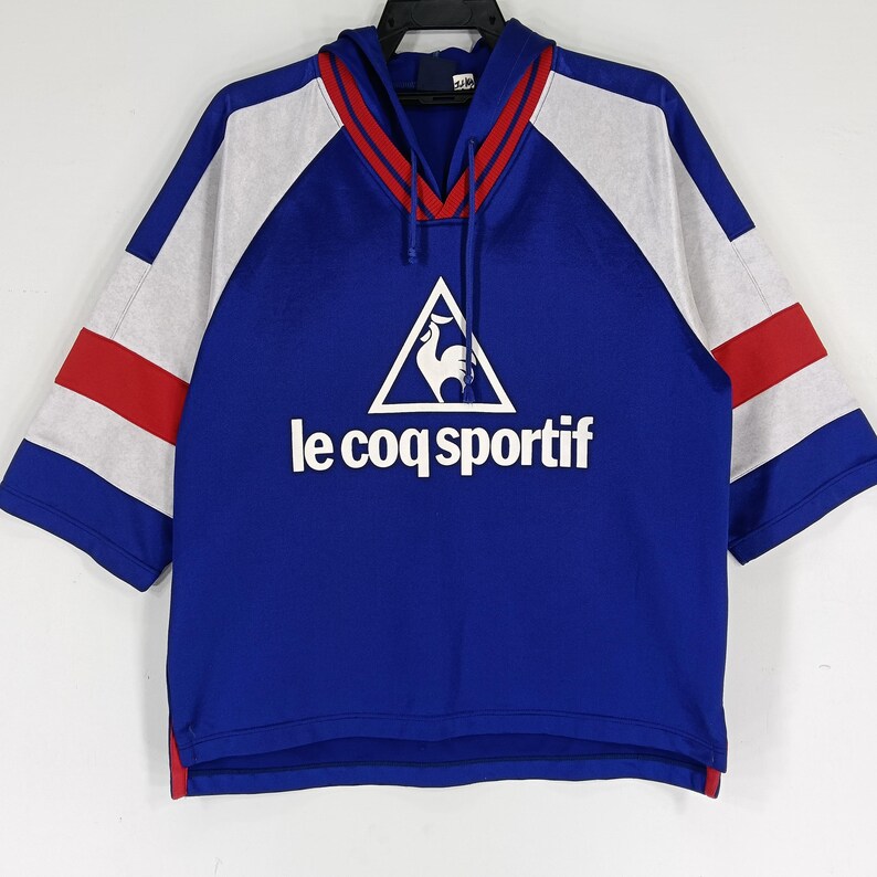 K&ouml;nnte beinhalten: Ein blaues, wei&szlig;es und rotes Kurzarm-Kapuzenpullover mit dem Le Coq Sportif Logo und einem Hahn-Design.