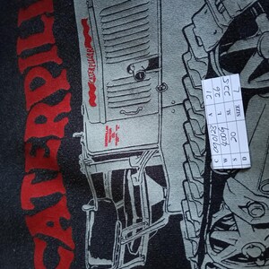 K&ouml;nnte beinhalten: Nahaufnahme eines schwarzen T-Shirts mit einer roten und grauen Grafik eines Caterpillar-Traktors. Das Wort "CATERPILLAR" ist vertikal in Rot gedruckt. Das T-Shirt hat ein Etikett der Gr&ouml;&szlig;e L. Ma&szlig;e: 21 cm, 26 cm, 22,5 cm.