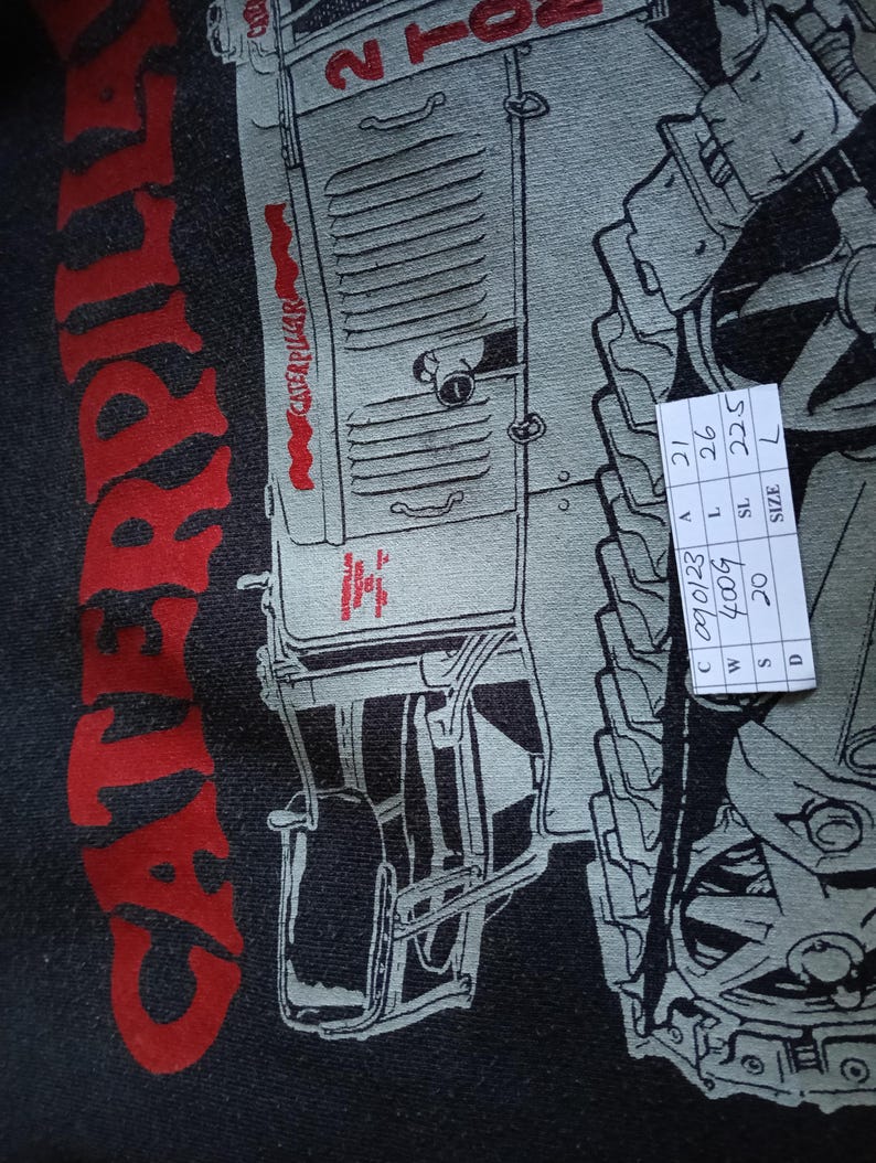 K&ouml;nnte beinhalten: Nahaufnahme eines schwarzen T-Shirts mit einer roten und grauen Grafik eines Caterpillar-Traktors. Das Wort "CATERPILLAR" ist vertikal in Rot gedruckt. Das T-Shirt hat ein Etikett der Gr&ouml;&szlig;e L. Ma&szlig;e: 21 cm, 26 cm, 22,5 cm.