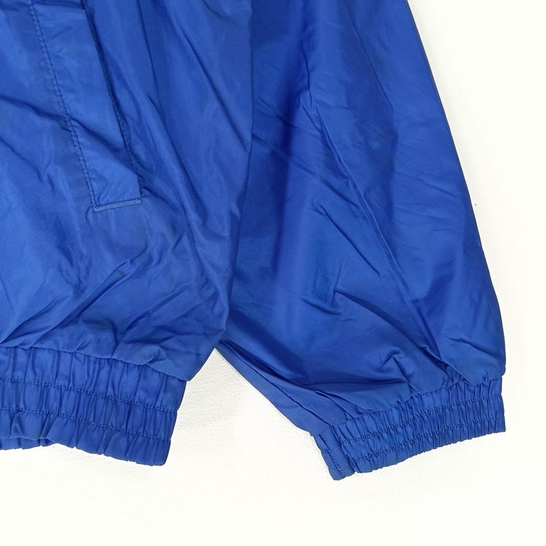 May include: Close-up of a vibrant blue jacket. The jacket features a gathered elastic cuff and hem, with a smooth, slightly shiny fabric. The design includes a pocket detail and a subtle sheen, suggesting a lightweight material.
