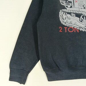 K&ouml;nnte beinhalten: Schwarzes Sweatshirt mit einer grauen Illustration eines Traktors und dem roten Text "2 TON". Das Sweatshirt hat lange &Auml;rmel und einen Rundhalsausschnitt. Das Design befindet sich auf der oberen rechten Seite.