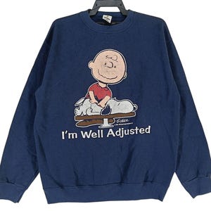 Peut inclure: Un sweat-shirt bleu marine avec une image de dessin animé de Charlie Brown et Snoopy. Le texte "I'm Well Adjusted" est imprimé sous l'image.