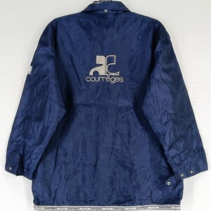 May include: A navy blue jacket with a white logo that says "Courreges" and a stylized "C" design. The jacket has a zipper closure and two pockets.