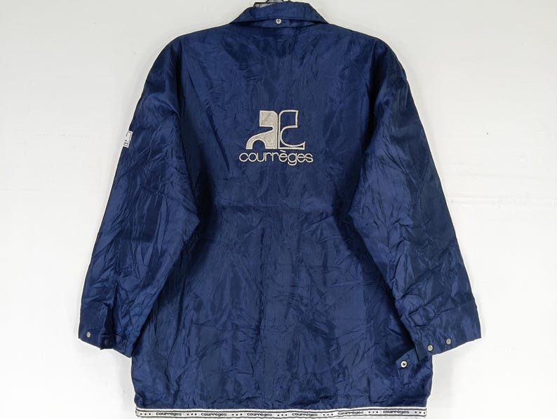 May include: A navy blue jacket with a white logo that says "Courreges" and a stylized "C" design. The jacket has a zipper closure and two pockets.