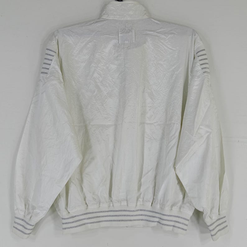May include: A white, vintage-style jacket with a high collar and a ribbed hem and cuffs. The jacket has a subtle, textured pattern and features gray stripes on the cuffs and sleeves. The back view of the jacket is shown.