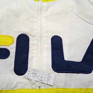 May include: White and yellow FILA jacket with a navy blue logo. The jacket has a full zipper and a tag with measurements. The jacket is a size L.