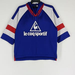 K&ouml;nnte beinhalten: Ein blaues, wei&szlig;es und rotes, kurz&auml;rmeliges Kapuzen-Sweatshirt mit dem Logo "le coq sportif" und einem Hahn in einem Dreieck.