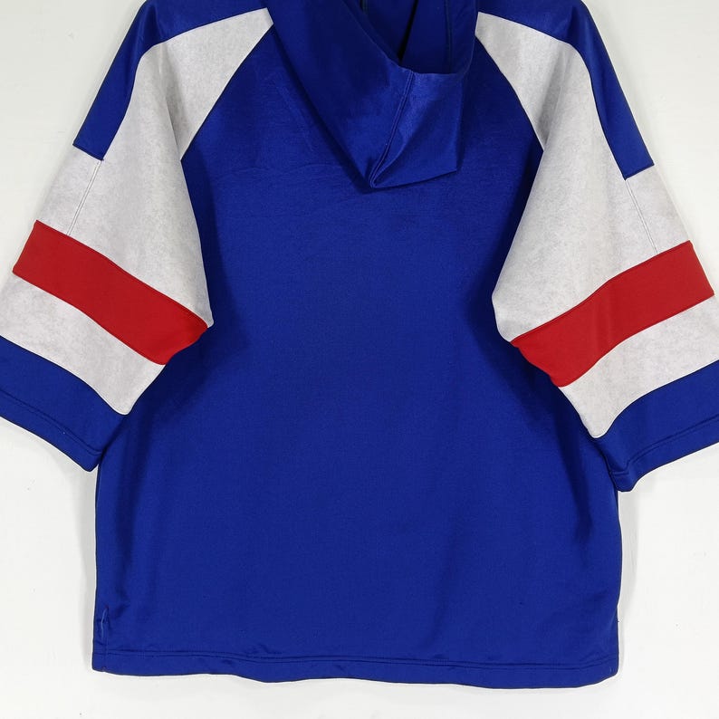 K&ouml;nnte beinhalten: Ein blaues Kapuzenpullover mit wei&szlig;en und roten Streifen&auml;rmeln. Der Pullover hat eine Kapuze mit Kordelzug und eine lockere Passform.