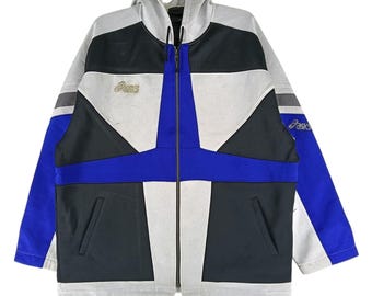 May include: A vintage athletic jacket in a colour-block design. The jacket features a white, black, and blue colour scheme with a full-length zip and a hood. The Asics logo is visible on the chest and sleeve. The jacket has a drawstring at the bottom.