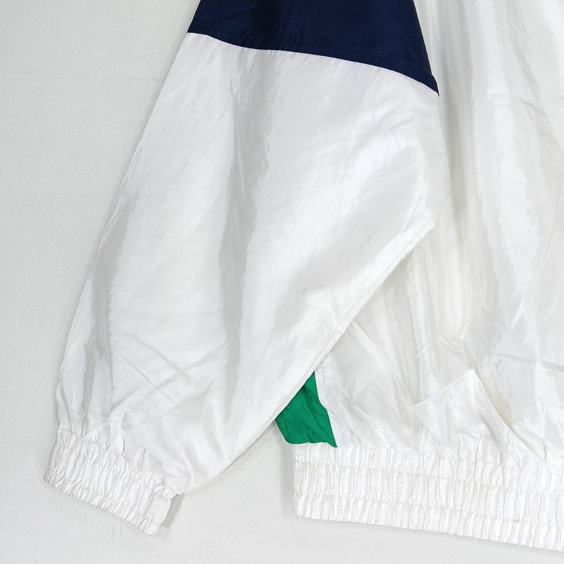 May include: White nylon jacket with navy blue and green accents. The jacket features a ribbed cuff and hem. The upper portion of the jacket is navy blue, with a white body and green accents.