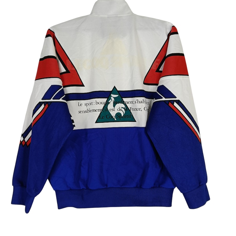 May include: A white, blue, and red jacket with a zipper closure. The jacket has a geometric pattern and a logo with the text "Le Coq Sportif" and the words "Le sport: bouger librement, s'habiller sensiblement est de la France, C".