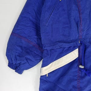 May include: Close-up of a blue and white vintage ski jacket. The jacket features a drawstring waist, a zippered pocket, and red stitching accents. The jacket has a white diagonal stripe and a blue drawstring.